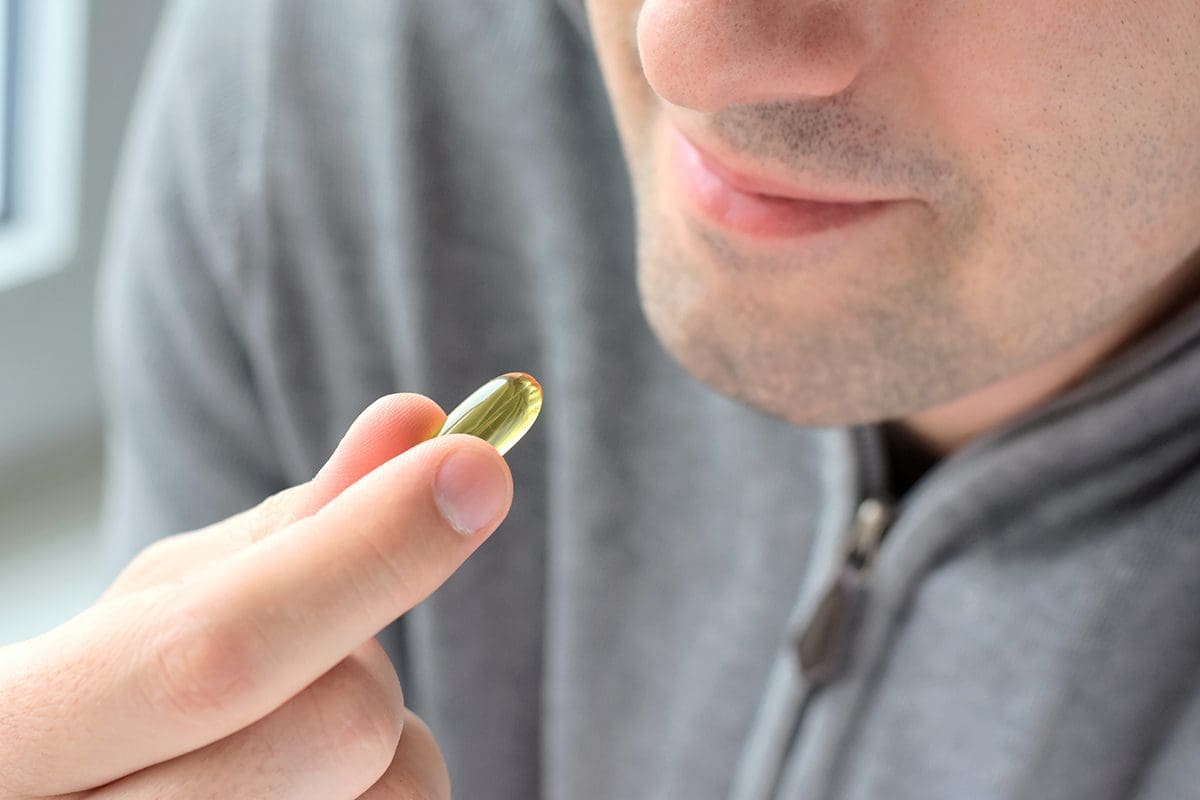Smiling man in a gray sweatshirt holding an Omega 3 soft gel in his fingers.